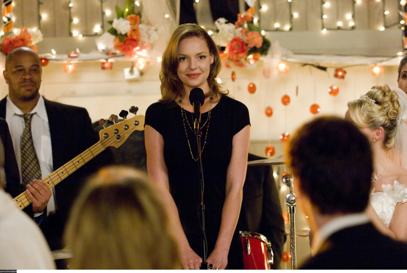 "27 sukienek" Kobieta w czarnej sukience stoi przy mikrofonie, z tyłu zespół muzyczny oraz dekoracje z kwiatów i światełek, po prawej w białej sukni panna młoda.