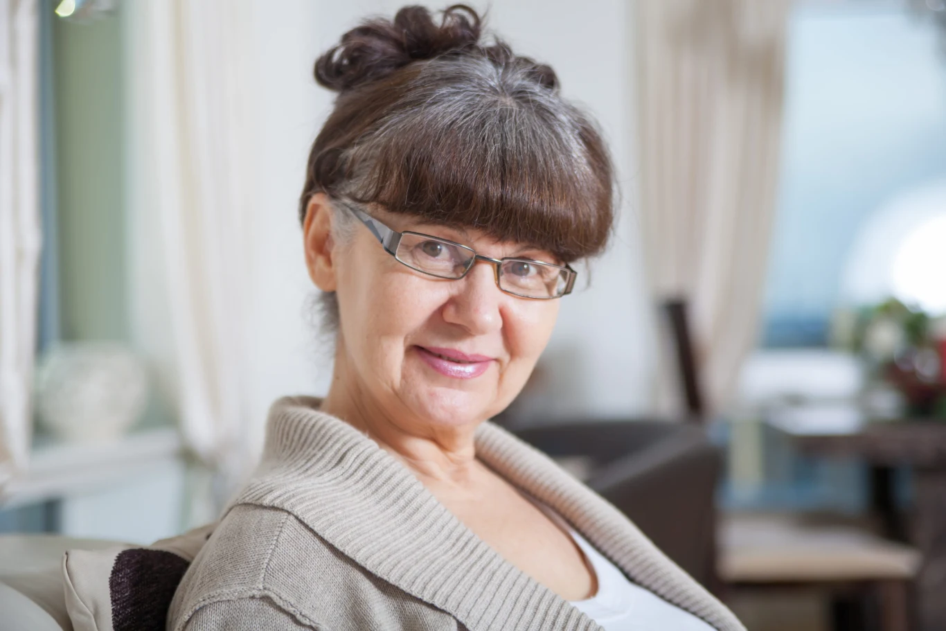 Starsza kobieta z okularami i spiętymi włosami, uśmiechnięta, ubrana w jasny sweter, siedzi w dobrze oświetlonym, przytulnym wnętrzu.