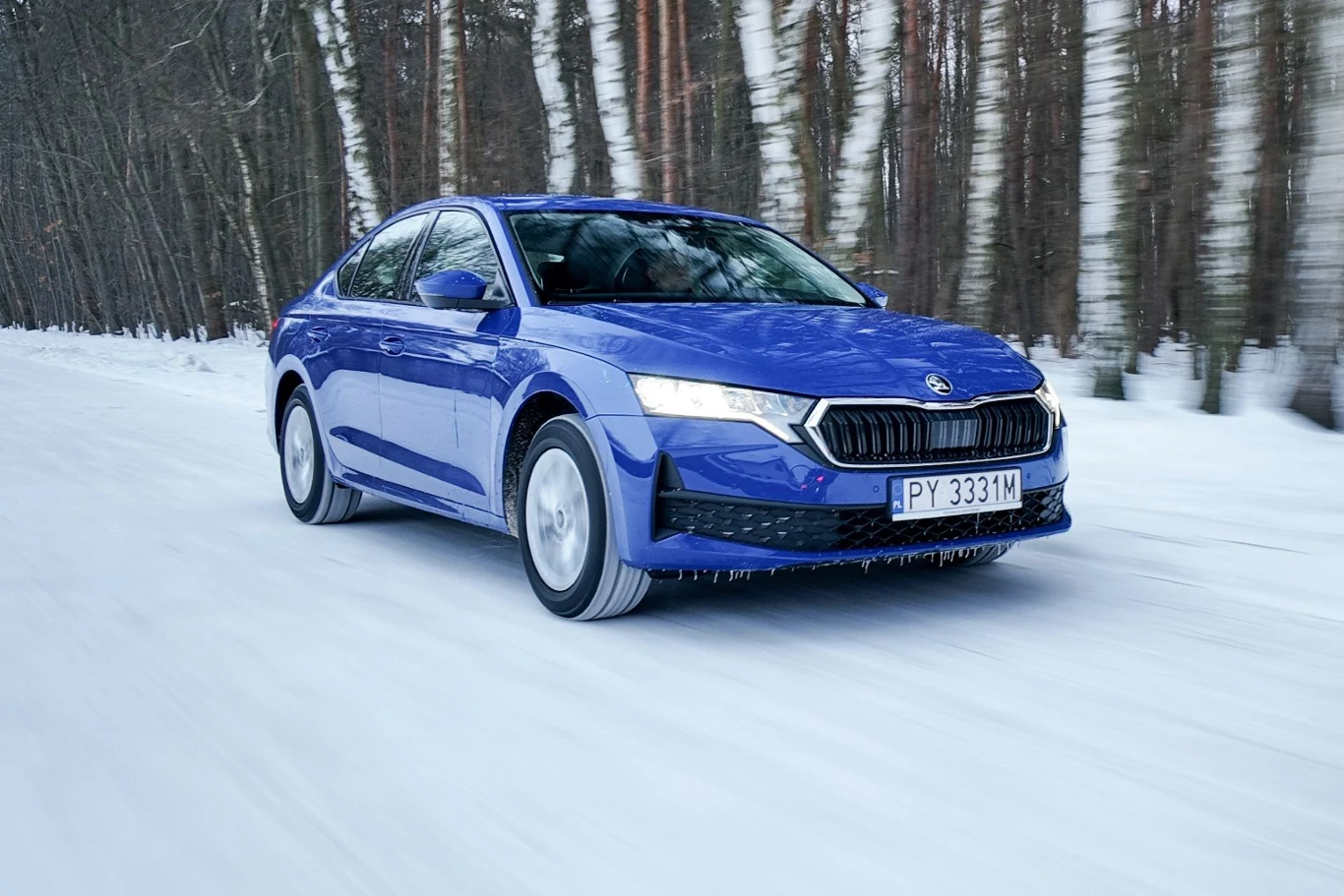 Niebieski sedan Skoda jedzie po ośnieżonej drodze otoczonej lasem z wysokimi, smukłymi drzewami.