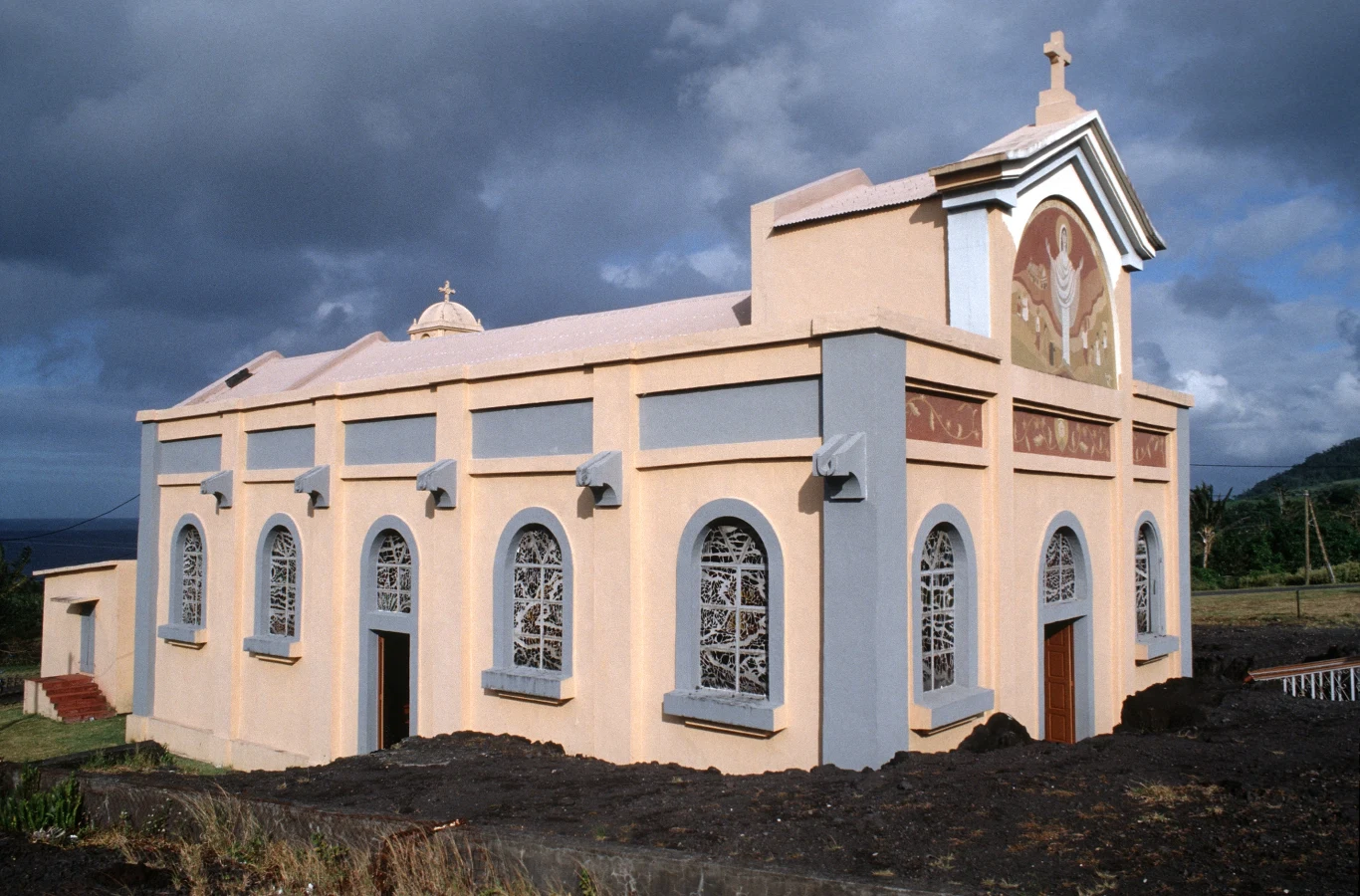Kościół o jasnobeżowych i szarych ścianach z dużymi, łukowatymi oknami, zdobiony freskiem z motywem religijnym nad głównym wejściem, otoczony ciemną, wulkaniczną ziemią, na tle pochmurnego nieba.