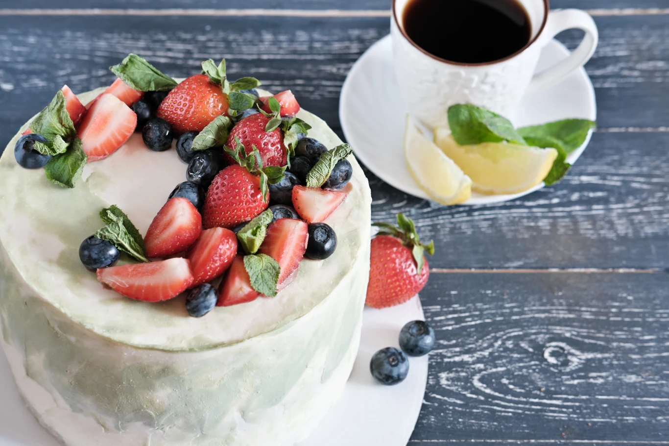 Wiosenny tort to świetny pomysł na deser na Wielkanoc. Sprawdzi się też na urodziny czy chrzciny. Tort z jasnym kremem, ozdobiony świeżymi owocami i listkami mięty, obok filiżanka czarnej kawy z cytryną na spodku.