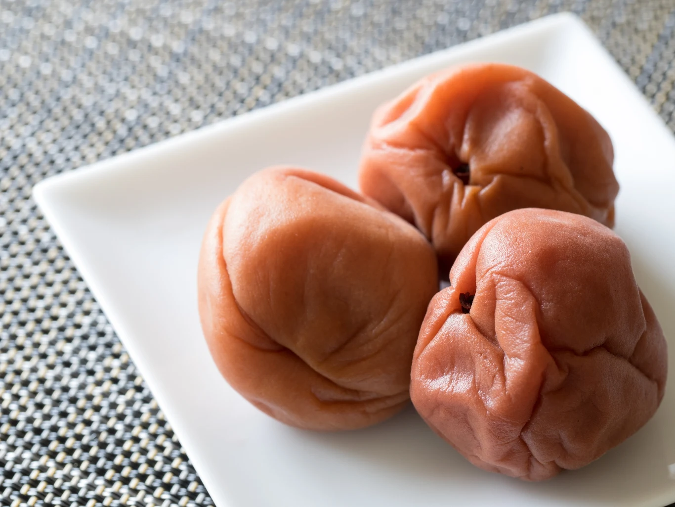 Trzy marynowane śliwki umeboshi o intensywnie pomarszczonej skórce leżące na białym kwadratowym talerzu.