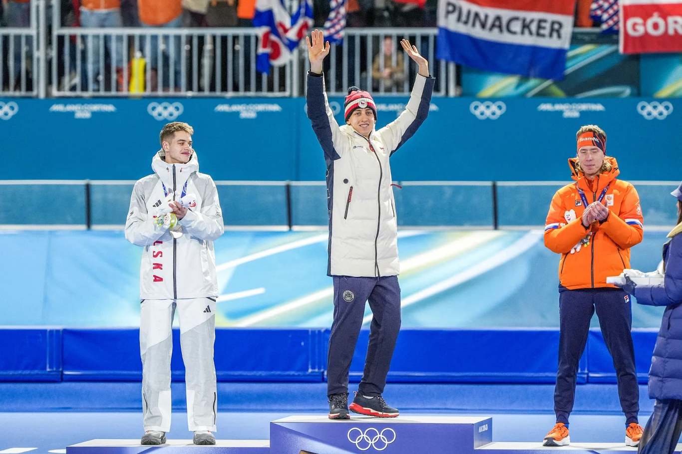 Zwycięzca w jasnej kurtce stoi na najwyższym stopniu podium z uniesionymi rękami, obok srebrny medalista w białym stroju oraz brązowy medalista w pomarańczowej kurtce, w tle flagi i kibice na trybunach.