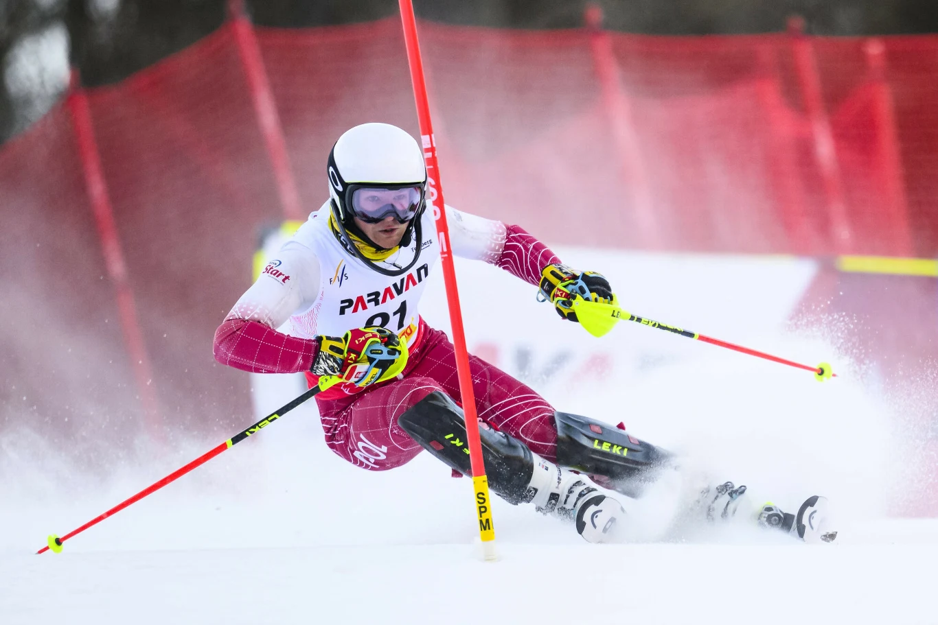 Narciarz w sportowym stroju z numerem startowym wykonuje dynamiczny skręt podczas slalomu na ośnieżonym stoku, mijając czerwony tyczkę i wzbijając śnieżny pył, w tle siatka zabezpieczająca trasę.