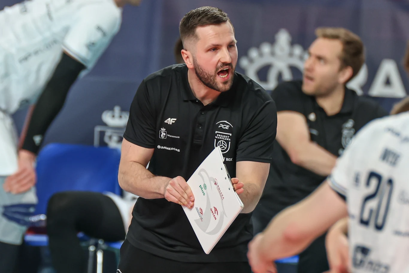 Trener drużyny siatkarskiej gestykuluje energicznie, trzymając w ręku tablicę taktyczną, przekazując wyraźne wskazówki zawodnikom podczas przerwy w meczu.