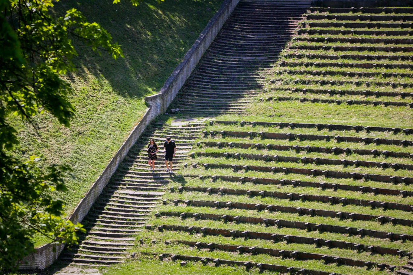 Dwoje ludzi spaceruje po szerokich, betonowych schodach otoczonych trawiastymi tarasami amfiteatru, oświetlonych słońcem i otoczonych zielenią.
