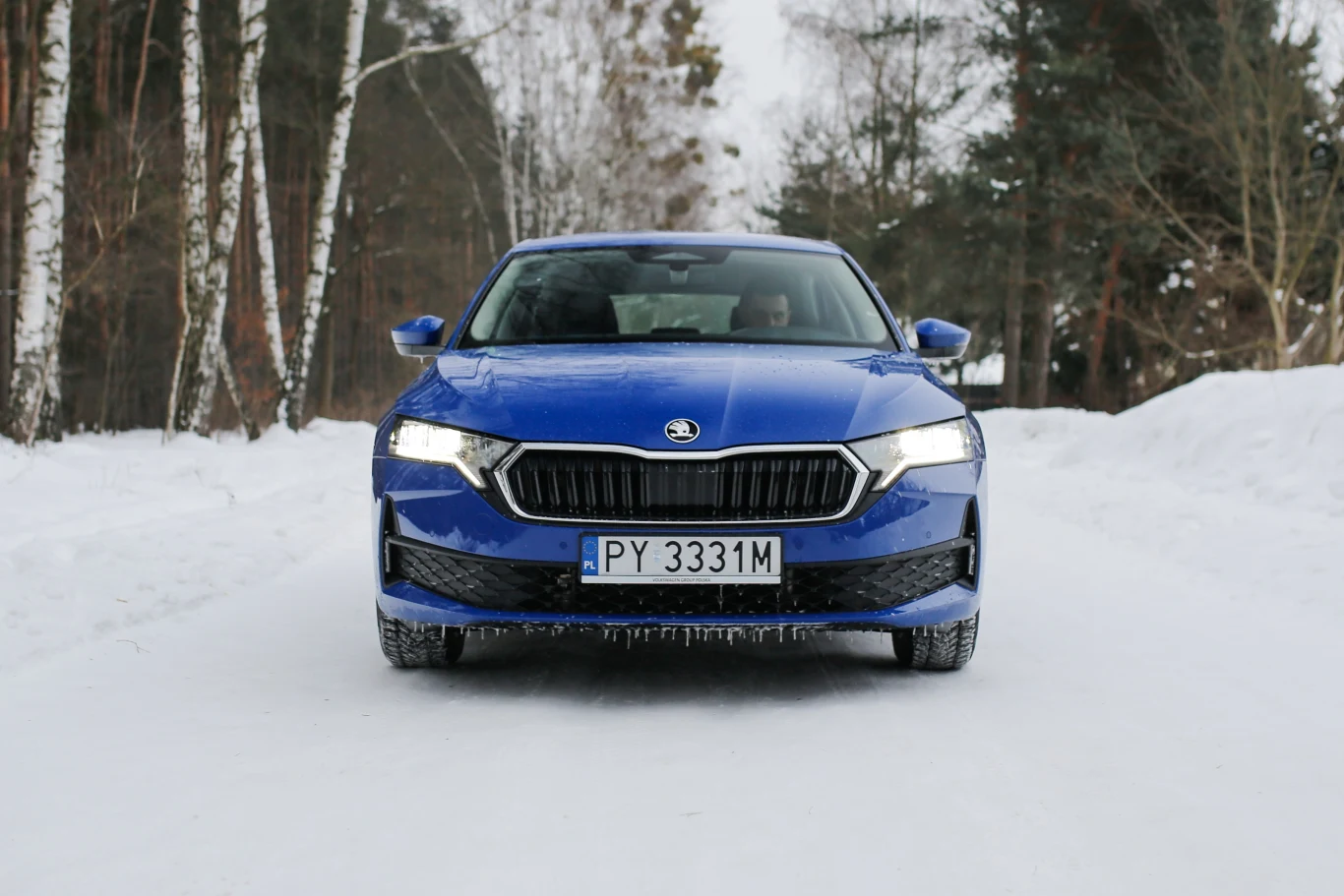 Niebieska Skoda Octavia z włączonymi światłami przednimi stoi na ośnieżonej drodze otoczonej drzewami bez liści.