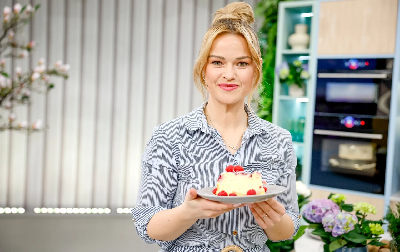 Uśmiechnięta kobieta trzyma elegancko udekorowany deser z malinami na białym talerzu, stojąc w nowoczesnej kuchni z kwiatami i piekarnikiem w tle.