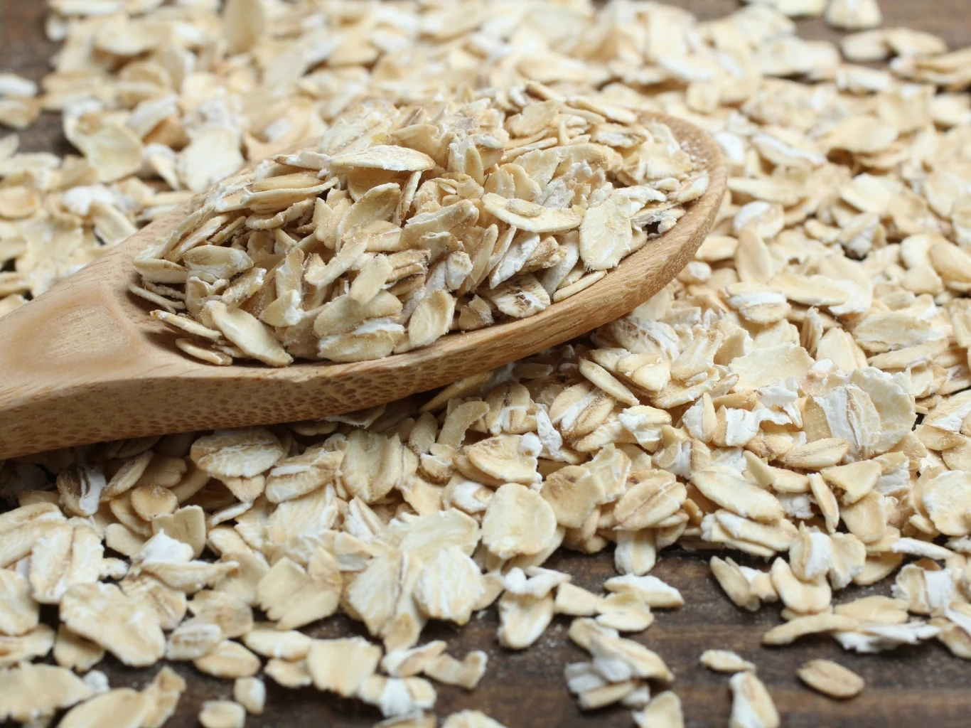 Drewniana łyżka wypełniona płatkami owsianymi leżąca na rozrzuconych płatkach owsianych. 