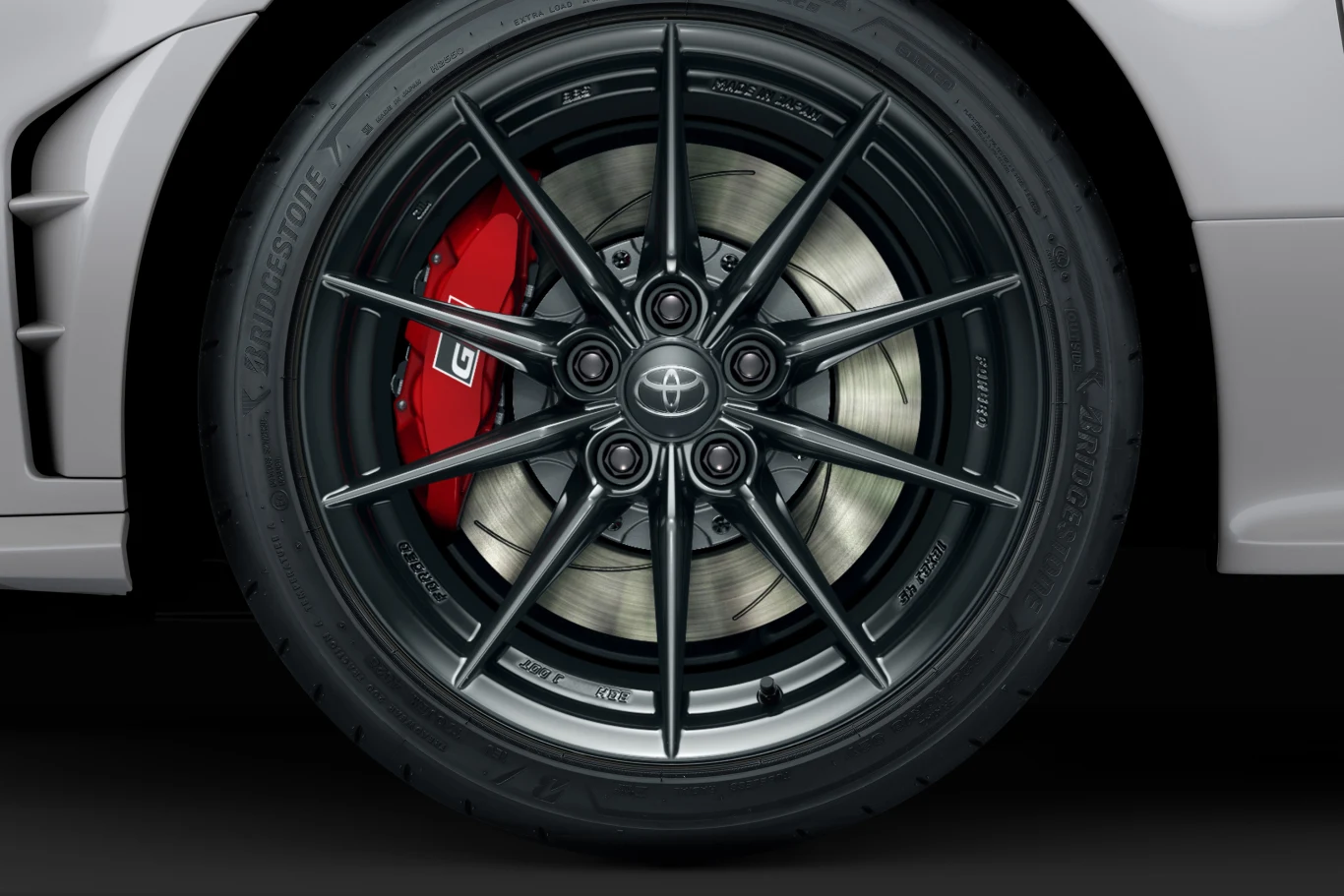 Toyota GR Yaris doczekała się kolejnych zmian. Co tym razem? Czarna, wieloramienna felga aluminiowa z widocznym czerwonym zaciskiem hamulcowym z logo GR oraz wentylowaną tarczą hamulcową, zamontowana na oponie Bridgestone przy nadkolu białego samochodu.