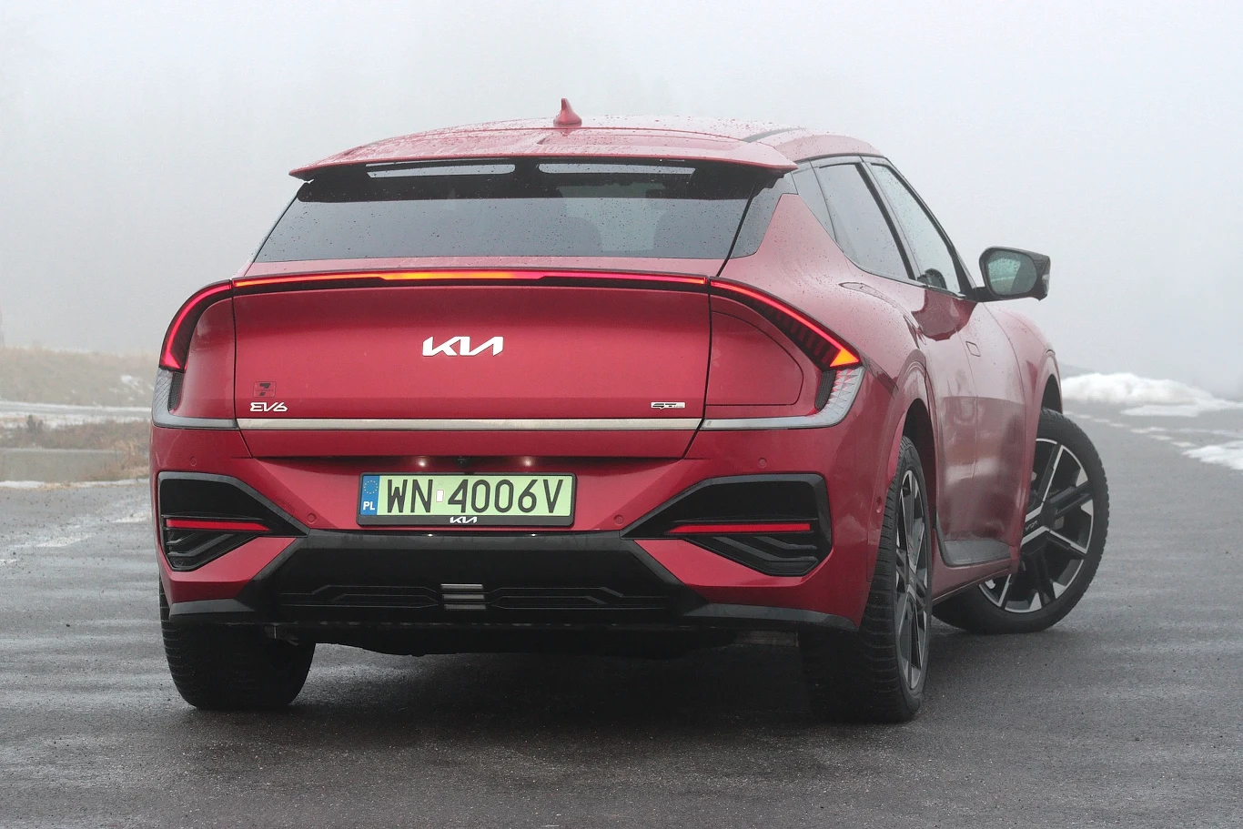 Czerwony samochód Kia EV6 ustawiony tyłem na mokrej nawierzchni, dobrze widoczne nowoczesne tylne lampy LED, aerodynamiczne linie nadwozia oraz charakterystyczne zielone tablice rejestracyjne wskazujące na pojazd elektryczny, w tle mglista i zamglona s...