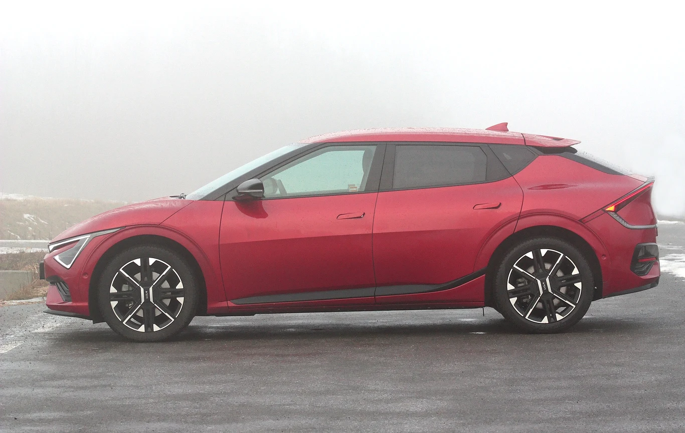 Czerwony, nowoczesny samochód elektryczny o dynamicznej sylwetce na ciemnej, mokrej nawierzchni, z dużymi sportowymi felgami i czarnymi detalami na drzwiach oraz z tyłu pojazdu, otoczony mgłą w tle.