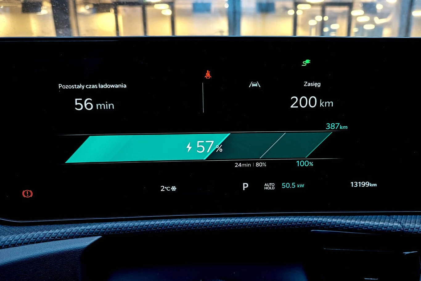 Cyfrowy wyświetlacz samochodu elektrycznego pokazujący poziom naładowania baterii na 57%, przewidywany zasięg 200 km, pozostały czas ładowania 56 minut, aktualną moc ładowania 50,5 kW oraz przebieg 13199 km.