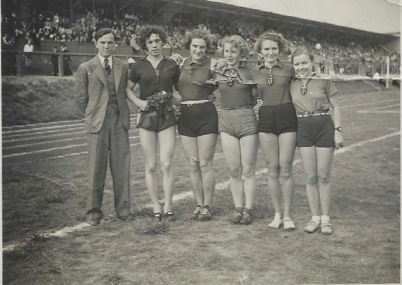 Grupa pięciu młodych sportsmenek ubranych w sportowe stroje i mężczyzna w garniturze stoją razem na bieżni stadionu lekkoatletycznego, w tle widoczna pełna kibiców trybuna.