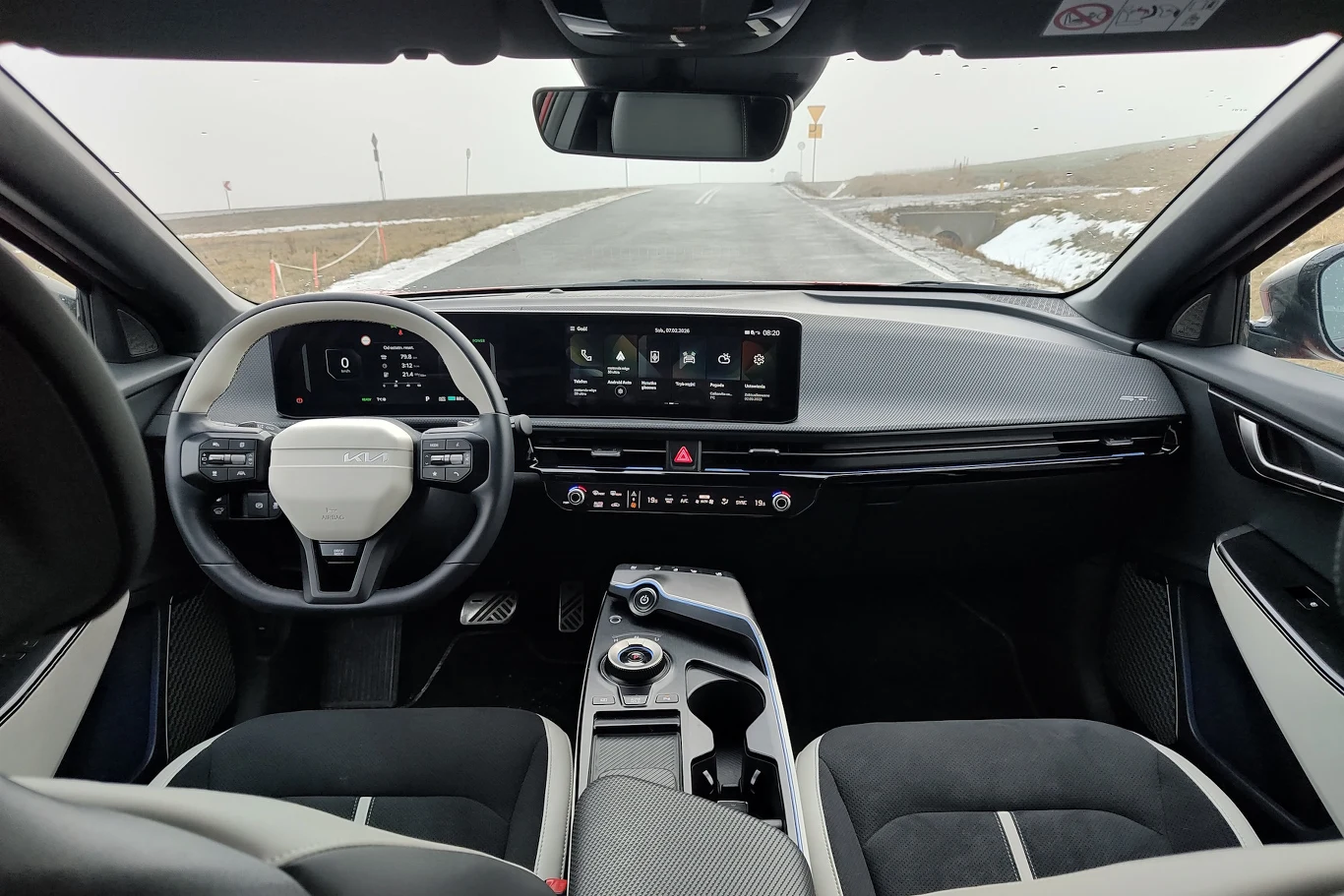 Nowoczesne wnętrze samochodu z cyfrowym zestawem wskaźników, dużym dotykowym ekranem centralnym, minimalistyczną deską rozdzielczą oraz panoramicznym widokiem przez przednią szybę na pustą drogę i zimowy krajobraz.