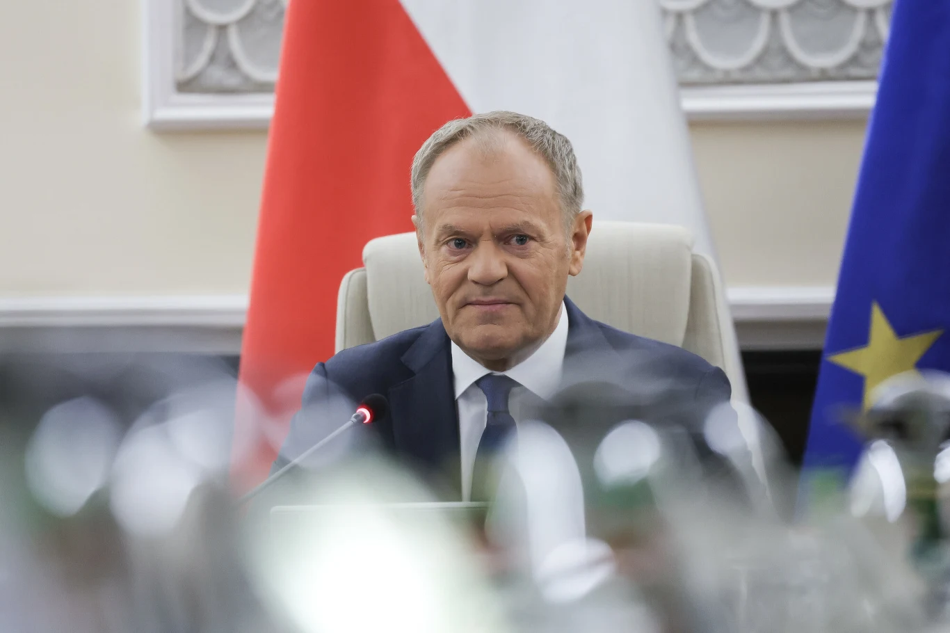 Mężczyzna w garniturze siedzi przy stole konferencyjnym, na tle flag Polski i Unii Europejskiej, przed nim stoi mikrofon.