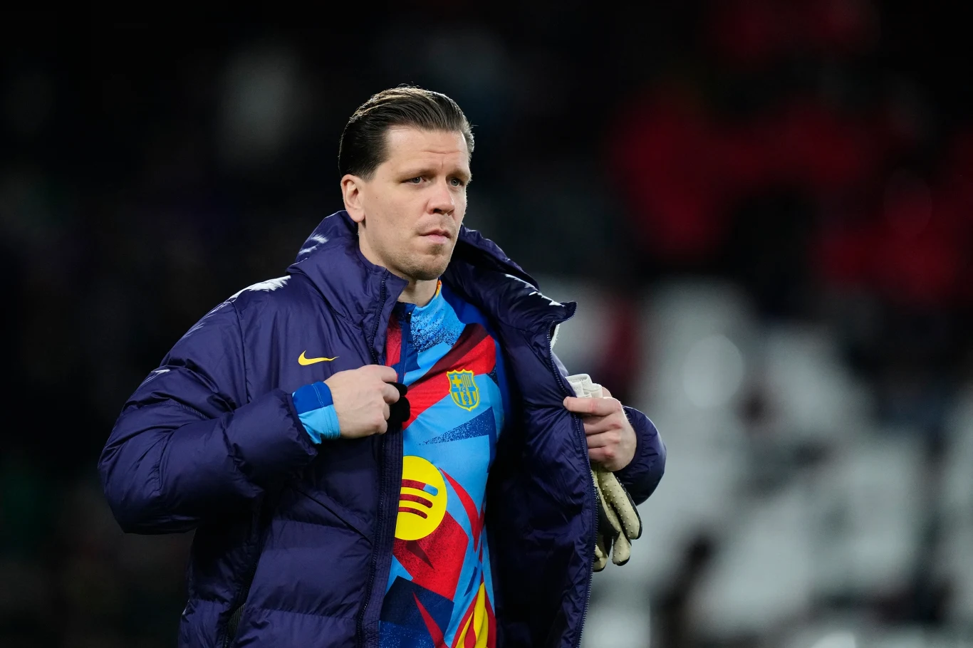 Mężczyzna w sportowej kurtce odsłania kolorową koszulkę z wyraźnym logo i wzorem na piersi, trzymając w dłoni jasne rękawice, na tle rozmytych trybun stadionu.