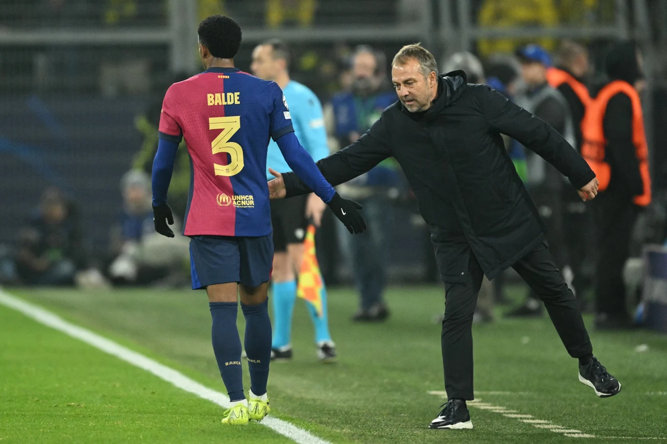 Piłkarz drużyny Barcelona w koszulce z numerem 3 opuszcza boisko, podczas gdy trener o blond włosach, ubrany na czarno, próbuje go zatrzymać lub przywołać, wyciągając rękę w jego kierunku.