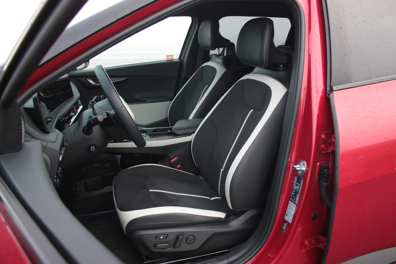Przestronne wnętrze samochodu z nowoczesnymi, czarno-białymi fotelami przednimi, widoczną deską rozdzielczą oraz kierownicą; czerwony lakier nadwozia kontrastuje z minimalistycznym designem kabiny.
