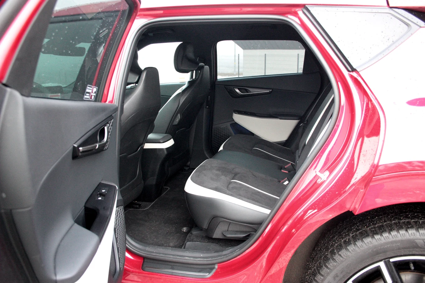 Wnętrze tylnej części samochodu z otwartymi drzwiami, czarne i szare skórzane siedzenia z kontrastującymi białymi akcentami, nowoczesne wykończenie, widoczne tylne koło oraz jasne elementy bocznej tapicerki.