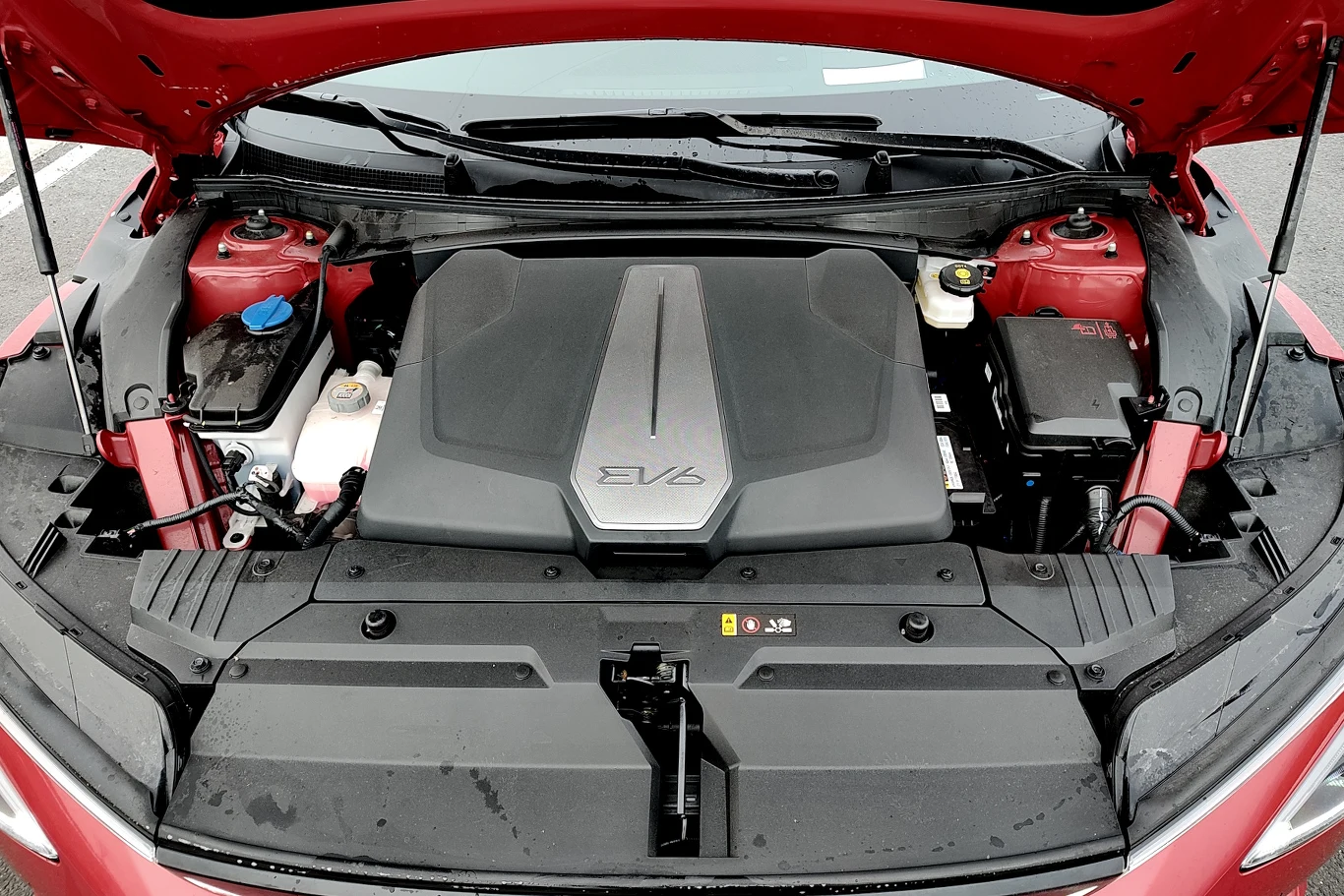Komora pod maską samochodu elektrycznego z logo EV6, centralnie umieszczoną pokrywą chroniącą komponenty elektryczne, widocznymi zbiornikami płynów eksploatacyjnych oraz solidnymi elementami konstrukcyjnymi po bokach.