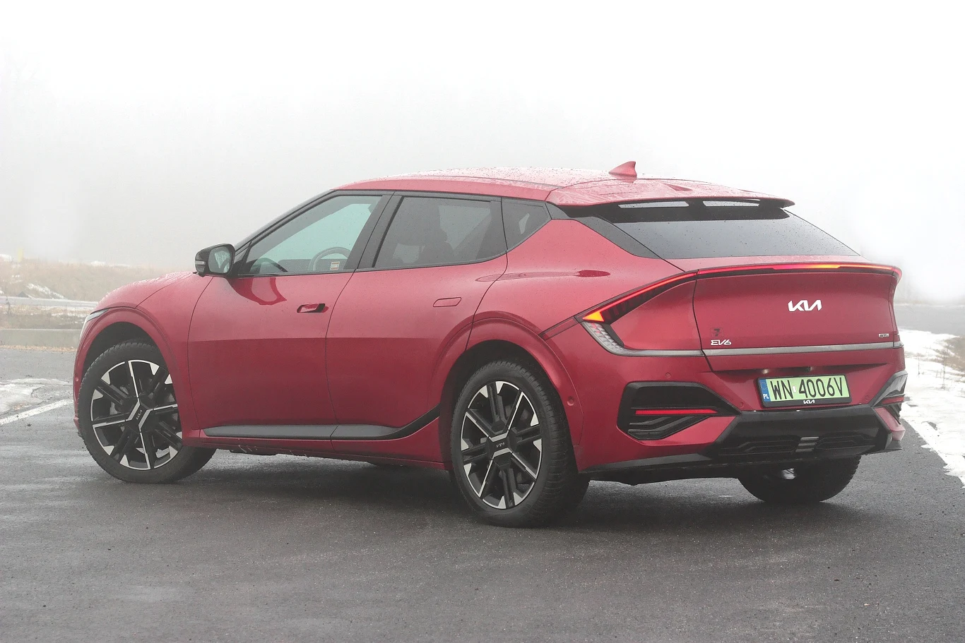 Czerwony samochód Kia EV6 ustawiony na mokrym asfalcie, z czarnymi detalami wokół nadkoli, aerodynamicznym tyłem i dużymi felgami, widoczna nowoczesna linia nadwozia oraz charakterystyczne światła LED z tyłu.