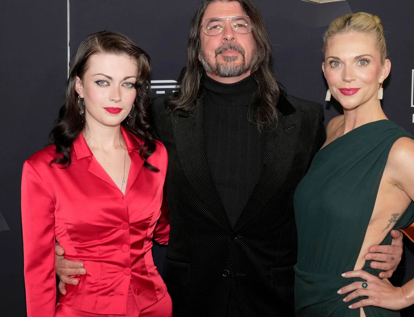 Troje elegancko ubranych osób pozuje wspólnie na tle ciemnej ścianki, kobieta po lewej ma na sobie czerwoną satynową sukienkę, mężczyzna w środku nosi czarny golf i okulary, a kobieta po prawej wybrała ciemnozieloną kreację na jedno ramię.