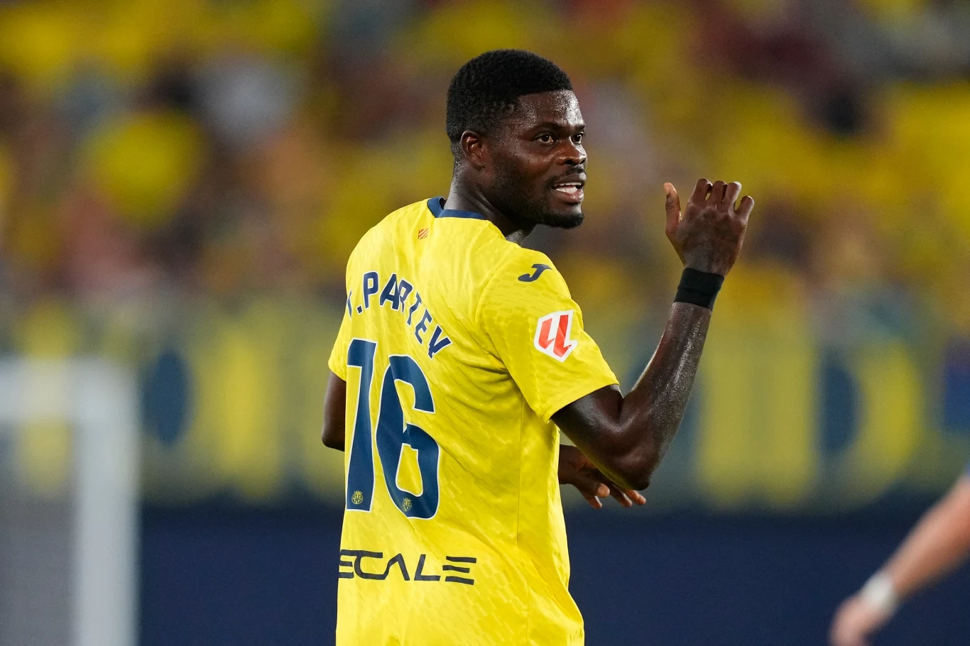 Piłkarz w żółtej koszulce z numerem 16 i napisem "P. PARTEY" odwrócony bokiem, machający ręką, na tle rozmytej, żółto-niebieskiej publiczności stadionowej.