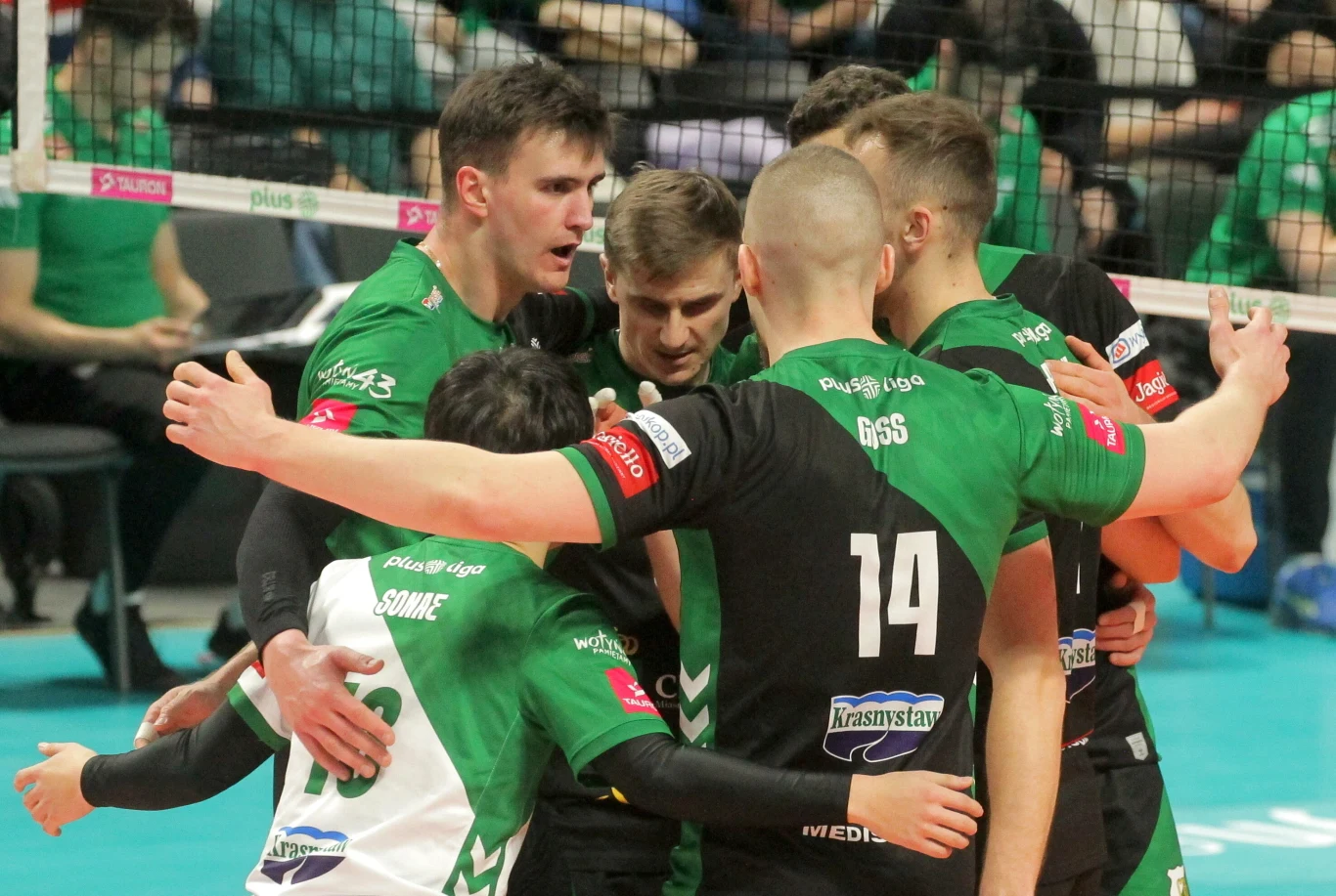 Grupa siatkarzy w zielono-czarnych strojach ściska się i motywuje w trakcie meczu, tworząc zwartą drużynę na boisku.