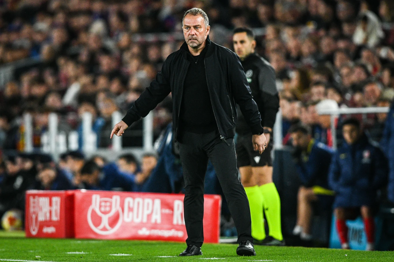 Trener w czarnej kurtce i spodniach stoi przy linii bocznej boiska piłkarskiego podczas meczu, w tle widoczny sędzia oraz trybuny wypełnione kibicami, a na pierwszym planie czerwona reklama turnieju Copa del Rey.