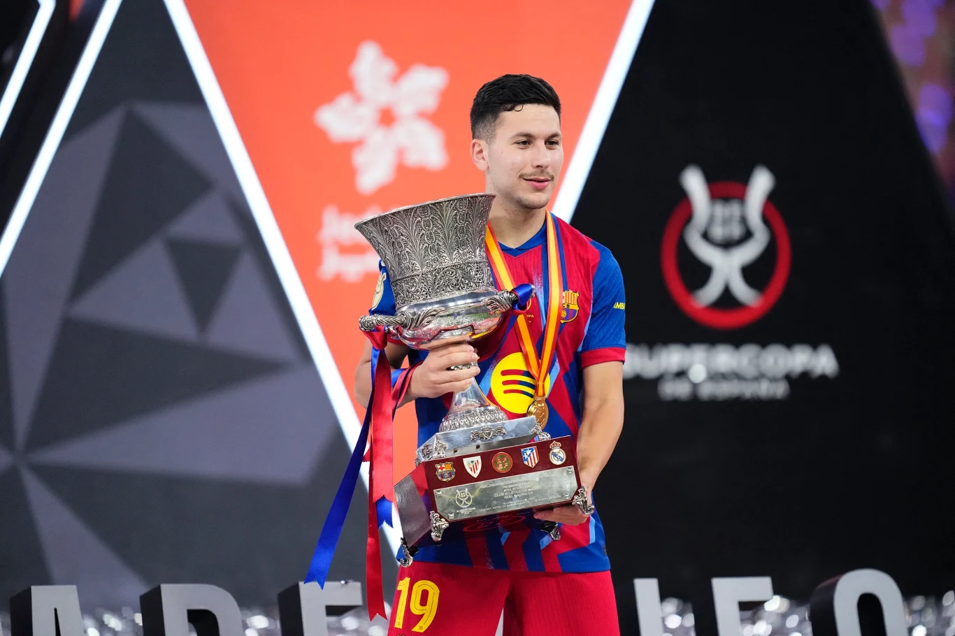 Młody sportowiec w stroju FC Barcelony trzyma okazały puchar ozdobiony wstążkami oraz medalem na szyi, stojąc na tle geometrycznego wzoru i symboli sportowych na arenie.