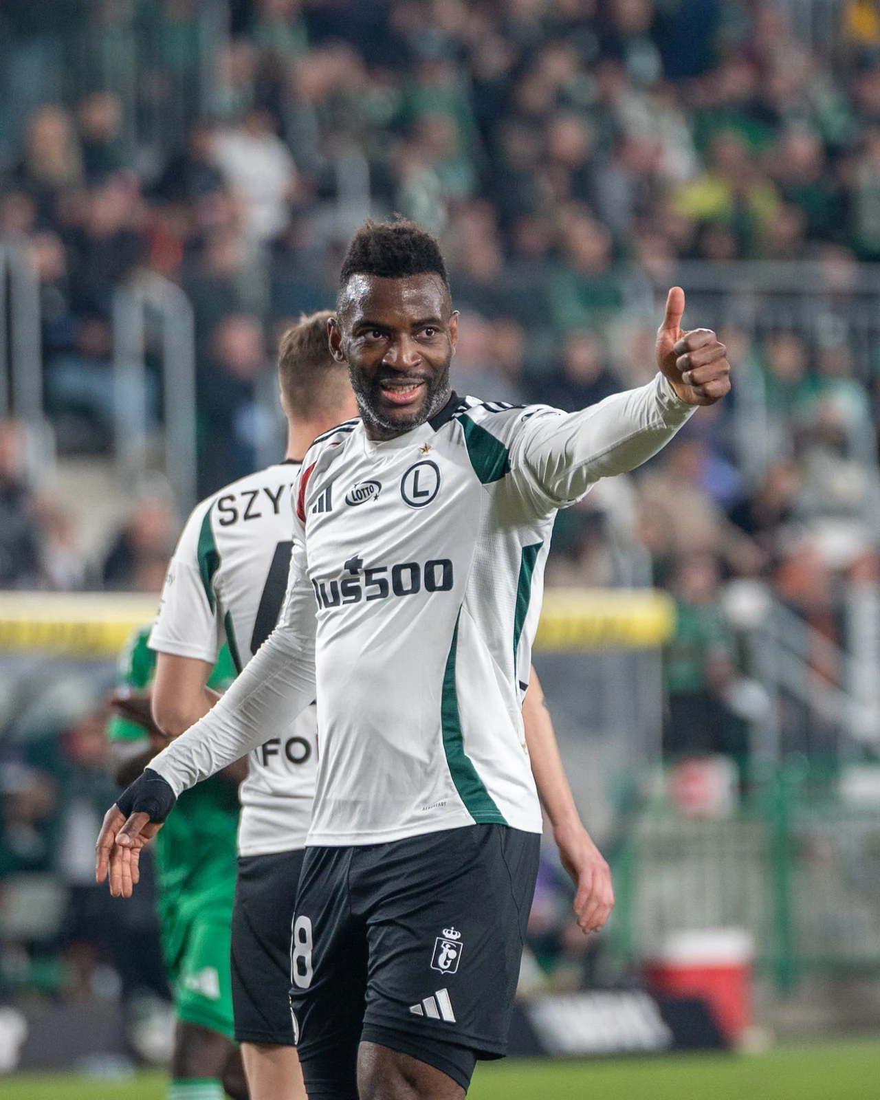 Piłkarz Legii Warszawa w białym stroju z numerem 8 okazuje radość, pokazując kciuk uniesiony w górę podczas meczu, w tle widoczni inni zawodnicy i rozmyte trybuny z kibicami.