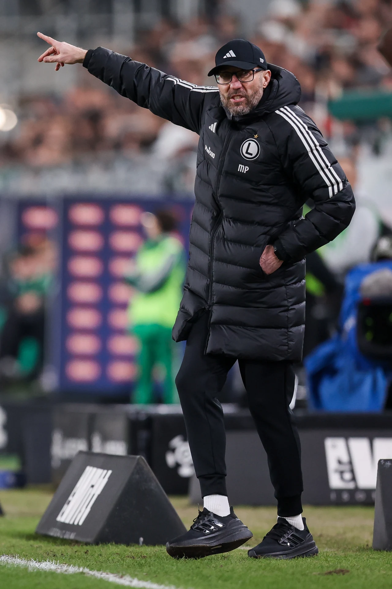 Trener drużyny Legii Warszawa ubrany w czarną puchową kurtkę z logo klubu i czapkę z daszkiem wydaje polecenia, wskazując kierunek ręką podczas meczu piłkarskiego na stadionie.