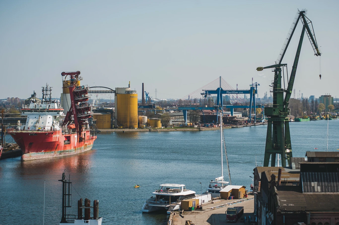 Port morski z dużym czerwonym statkiem na wodzie, charakterystycznymi żółtymi zbiornikami oraz wysokimi przemysłowymi dźwigami w tle.