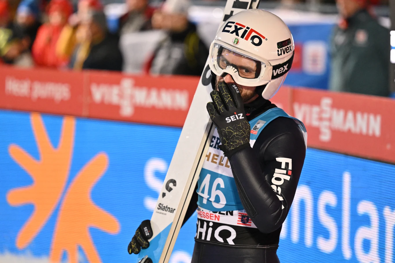 Skoczek narciarski w kombinezonie i kasku trzyma narty w jednej ręce, a drugą zasłania usta; w tle widoczne rozmazane logo Viessmann i fragment kolorowego baneru zawodów.