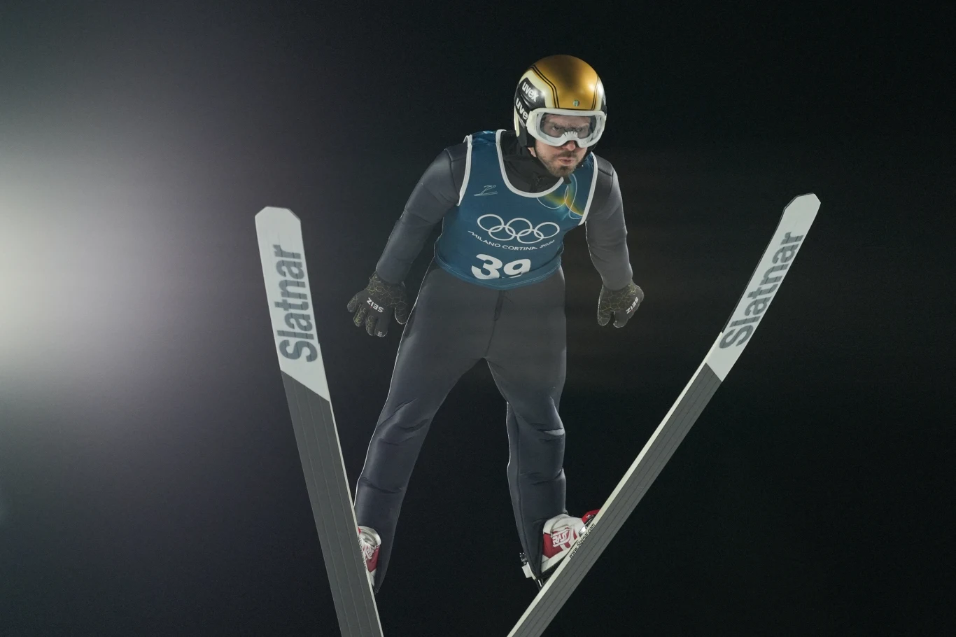 Skoczek narciarski w stroju sportowym leci w powietrzu z szeroko rozstawionymi nartami, na głowie ma złoty kask i gogle, na piersi widoczny numer startowy oraz logo igrzysk olimpijskich.