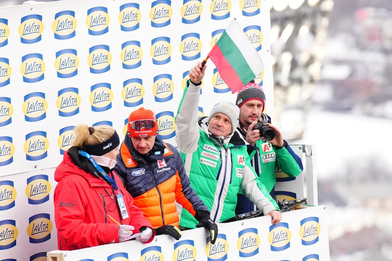 Czterech ludzi ubranych w zimowe kurtki stoi na podium z tłem logo sponsora; jeden z nich trzyma bułgarską flagę, inny fotografuje, pozostali są skoncentrowani na obserwacji wydarzenia.