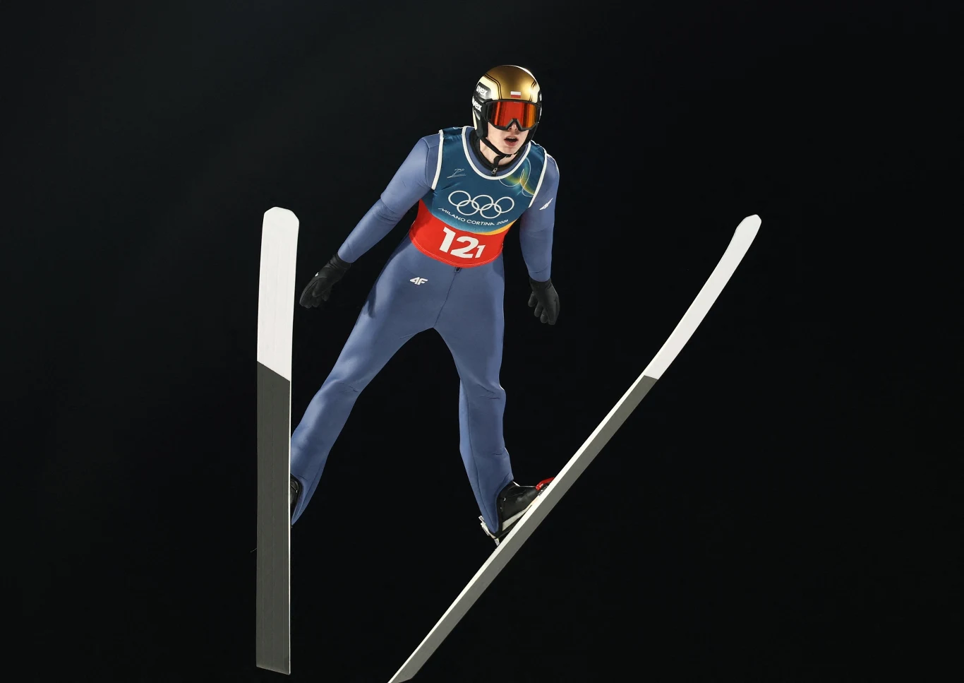 Skoczek narciarski w niebieskim kombinezonie i hełmie w locie, z widocznym numerem startowym 121 oraz symbolami olimpijskimi, na tle czarnego tła.