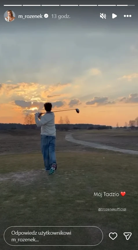 Chłopiec w jasnym swetrze i niebieskich spodniach trzyma kij golfowy na tle zachodzącego słońca na polu golfowym.