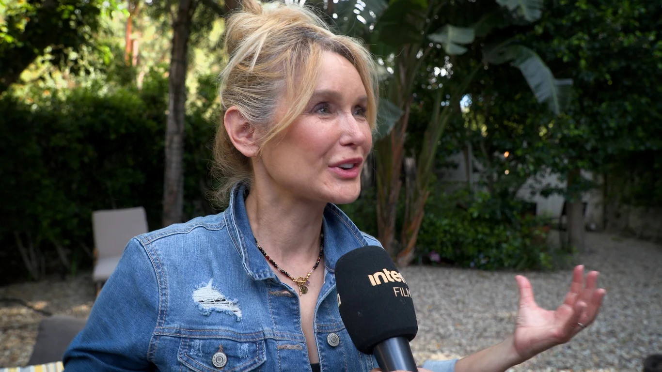 Katarzyna Śmiechowicz Kobieta w dżinsowej kurtce z podniesioną ręką udziela wywiadu, trzymając mikrofon z logo 'interFILM', w tle widoczne drzewa i roślinność ogrodowa.