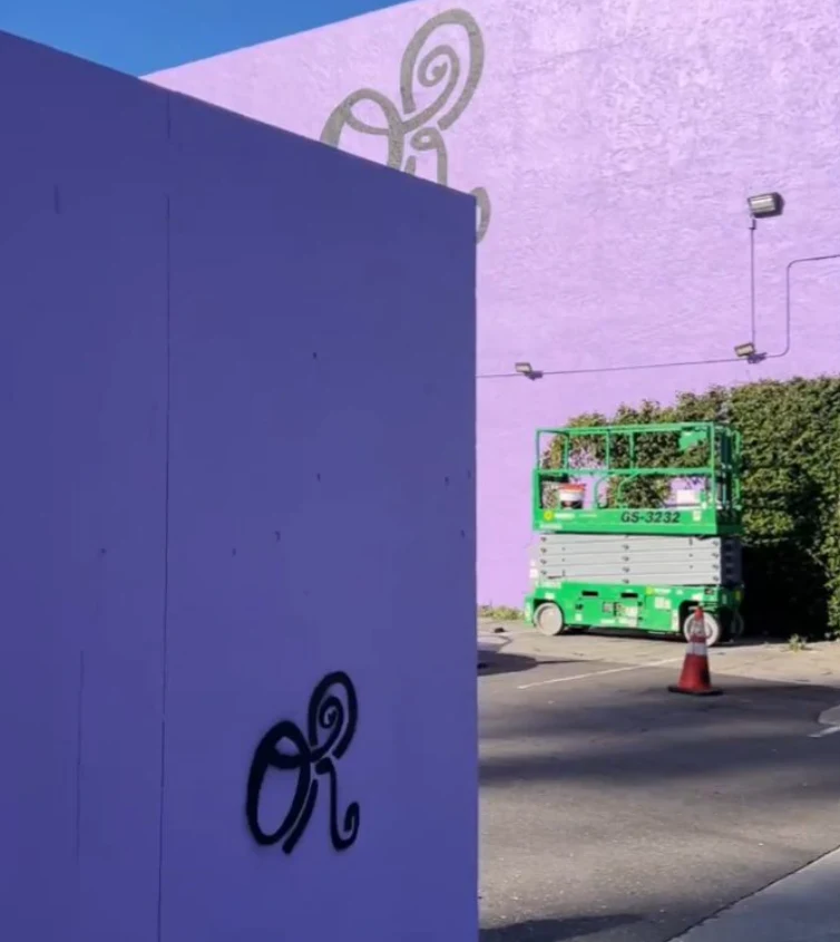 Duża zielona podnośnik koszowy stoi na tle fioletowej ściany ozdobionej spiralnym wzorem; na pierwszym planie znajduje się fioletowy panel z czarnym graffiti w stylu spirali, na ziemi ustawiony jest pomarańczowy pachołek ostrzegawczy.