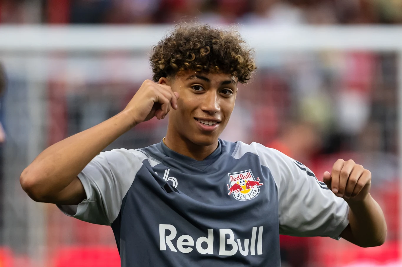 Młody piłkarz w szarej koszulce Red Bull Salzburg wykonuje gesty rękami, uśmiechając się szeroko, na tle zamazanych trybun stadionu.