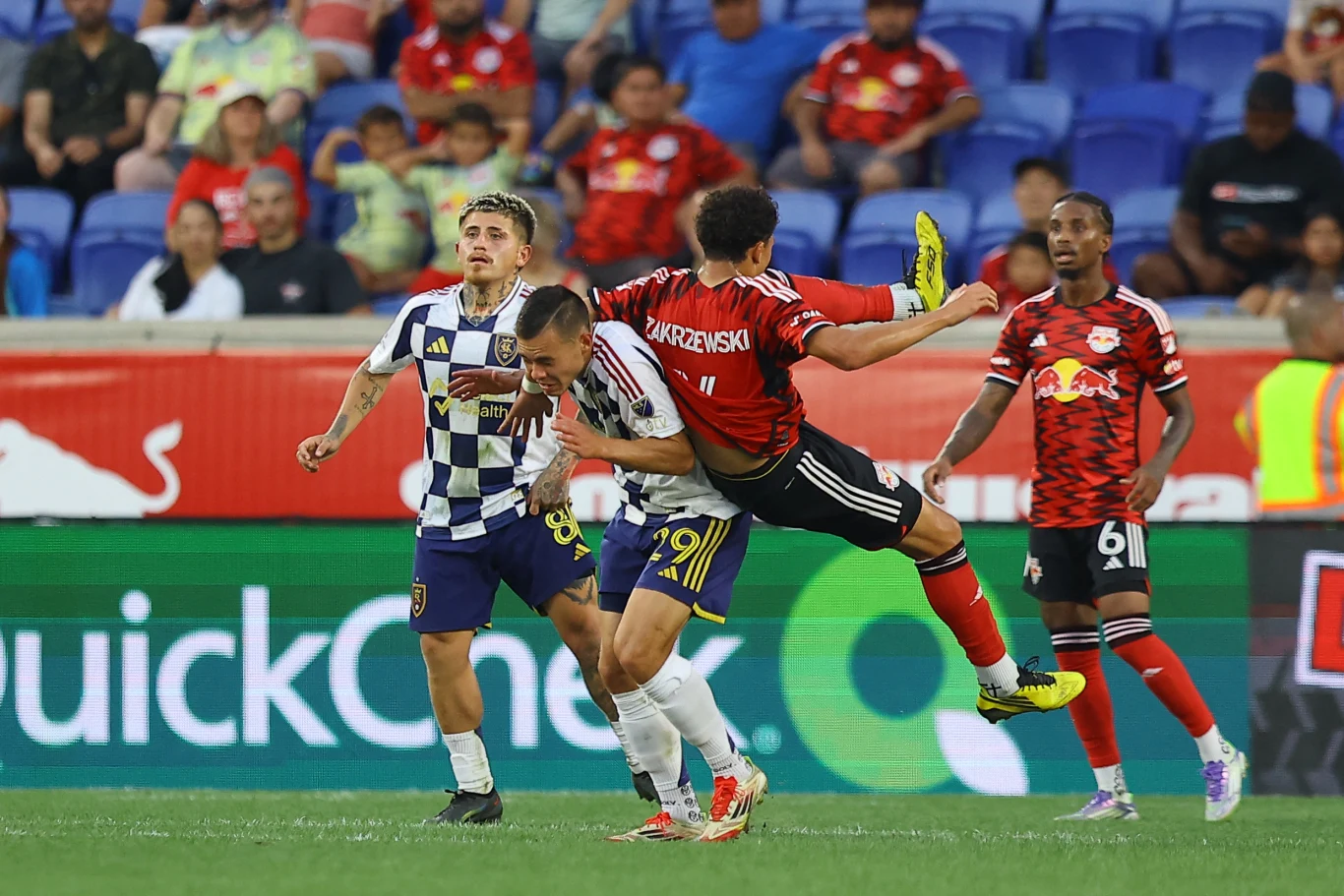 Zawodnik w czerwono-czarnej koszulce wykonuje dynamiczny wślizg, zderzając się z piłkarzem drużyny w białych i granatowych barwach, podczas gdy pozostali gracze obserwują sytuację na tle pustych niebieskich trybun.