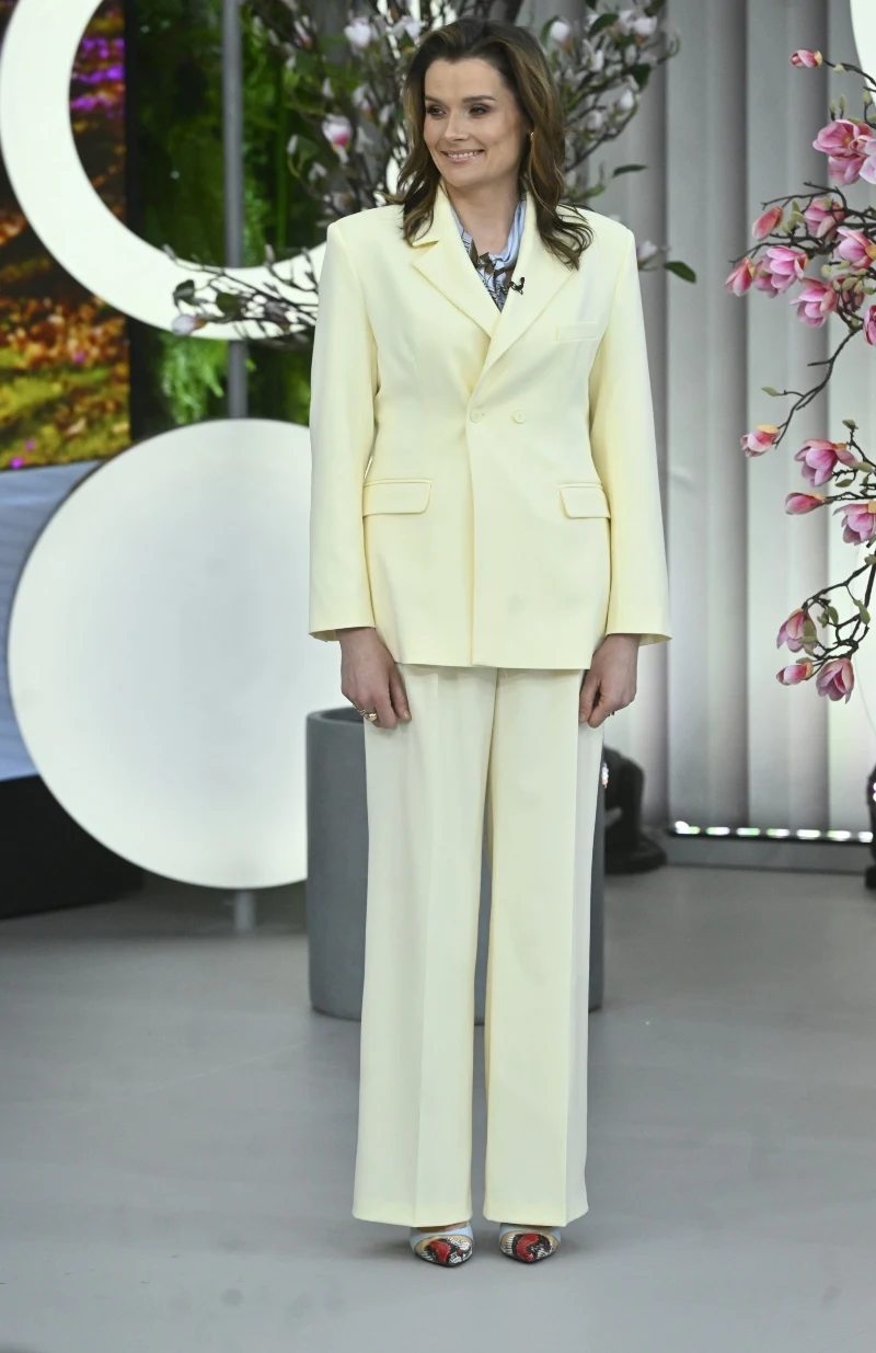 Kobieta w kremowym garniturze i kolorowych butach na obcasie stoi na tle aranżacji z kwitnącymi różowymi gałązkami, w jasnym, nowoczesnym wnętrzu.