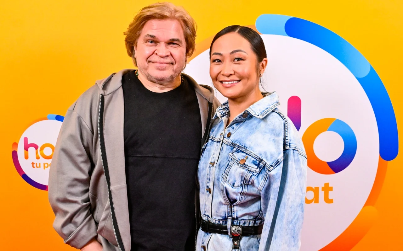 Dwie uśmiechnięte osoby stojące obok siebie na tle kolorowej grafiki z napisem, mężczyzna w szarej kurtce i czarnej koszulce, kobieta w jasnej dżinsowej kurtce z ciemnymi włosami upiętymi do tyłu.