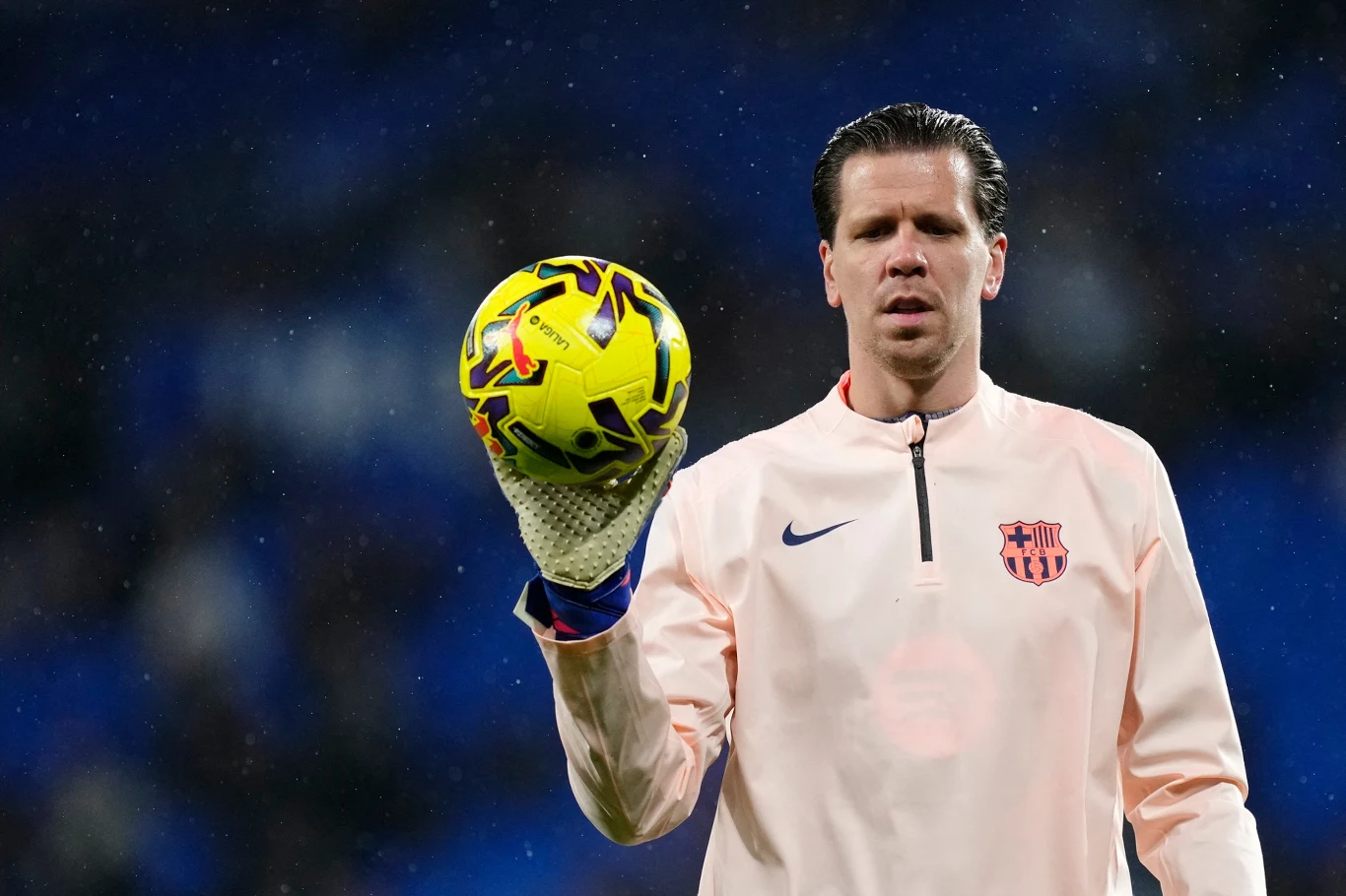 Bramkarz w jasnej bluzie z logo FC Barcelony trzyma w jednej dłoni piłkę nożną, skupiony patrząc przed siebie, na tle rozmytego, ciemnoniebieskiego stadionu.