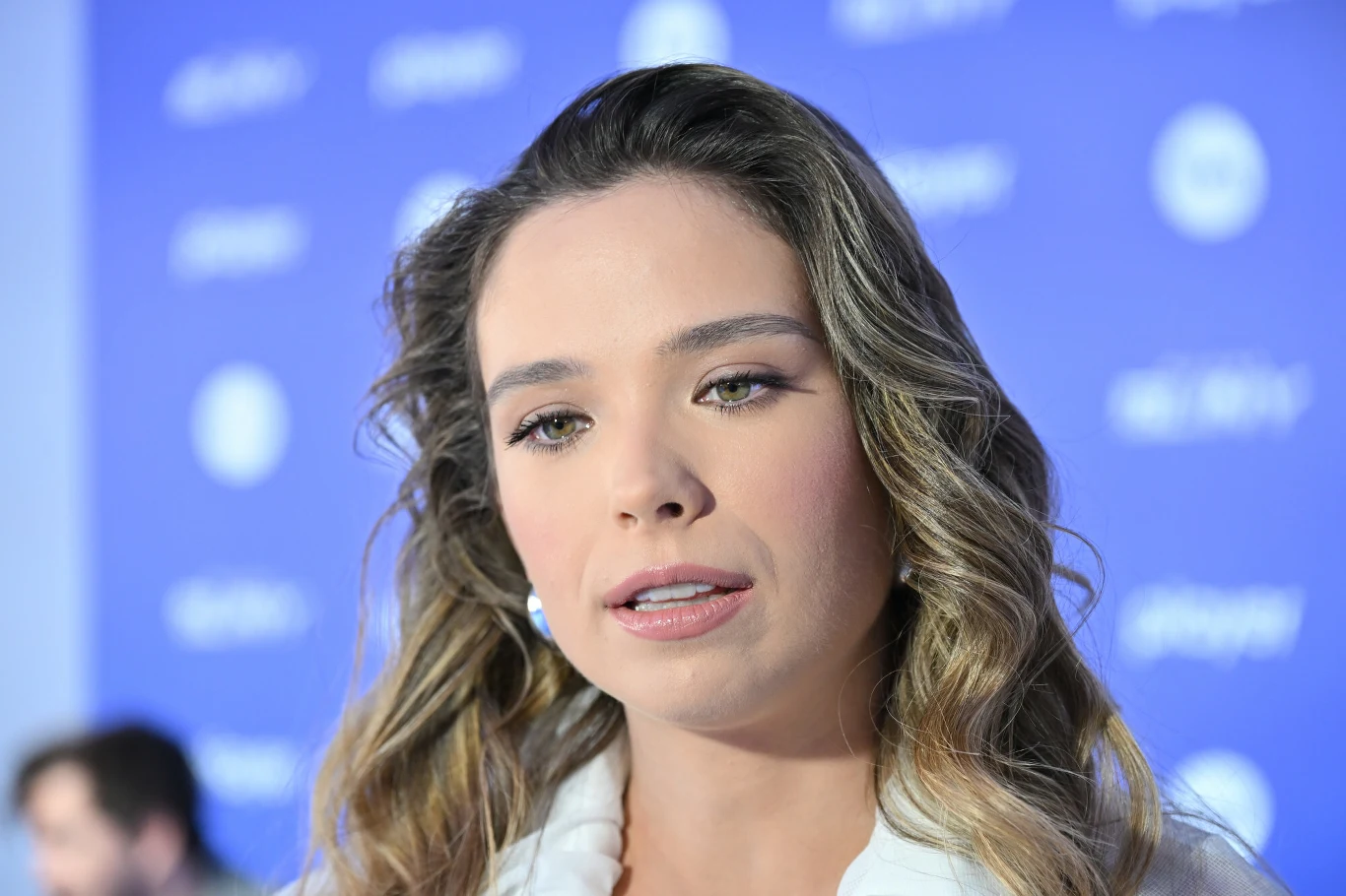 Młoda kobieta z długimi, falującymi włosami w jasnym stroju stoi na tle niebieskiej ścianki z drobnymi, rozmazanymi napisami i logotypami, patrząc lekko w bok ze spokojnym wyrazem twarzy.