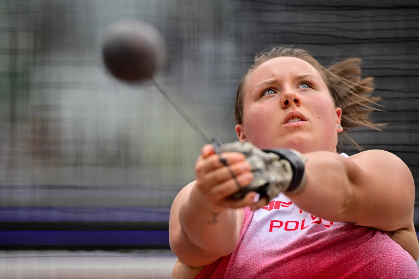 Zawodniczka w sportowej koszulce z napisem „Polska” dynamicznie wykonuje rzut młotem, skupiona na wysiłku i precyzji ruchu, młot znajduje się w ruchu tuż przed wypuszczeniem.