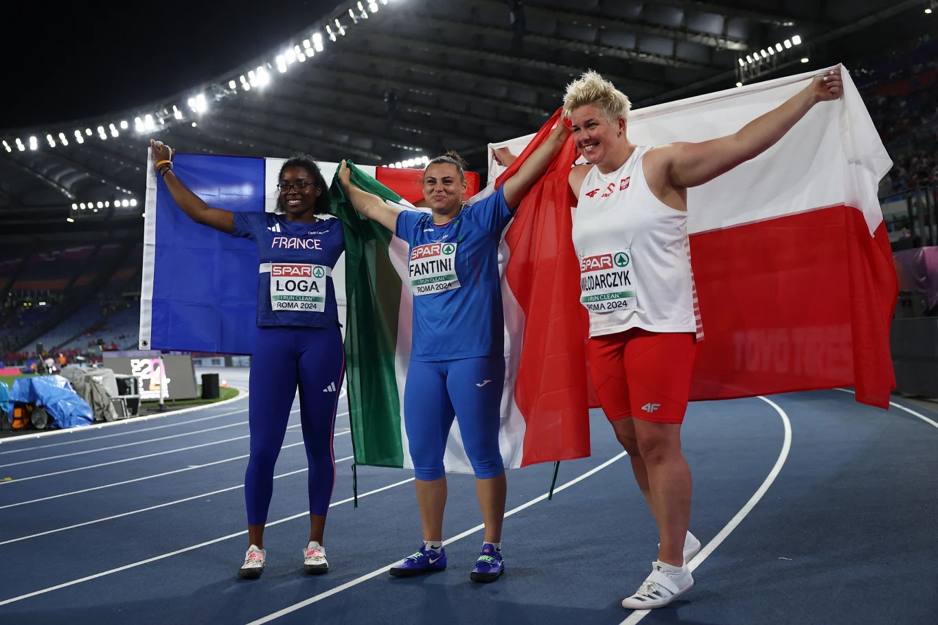 Trzy sportsmenki stoją na bieżni stadionu lekkoatletycznego z szeroko rozpostartymi flagami Francji, Włoch i Polski, wyrażając radość po zakończonych zawodach sportowych.