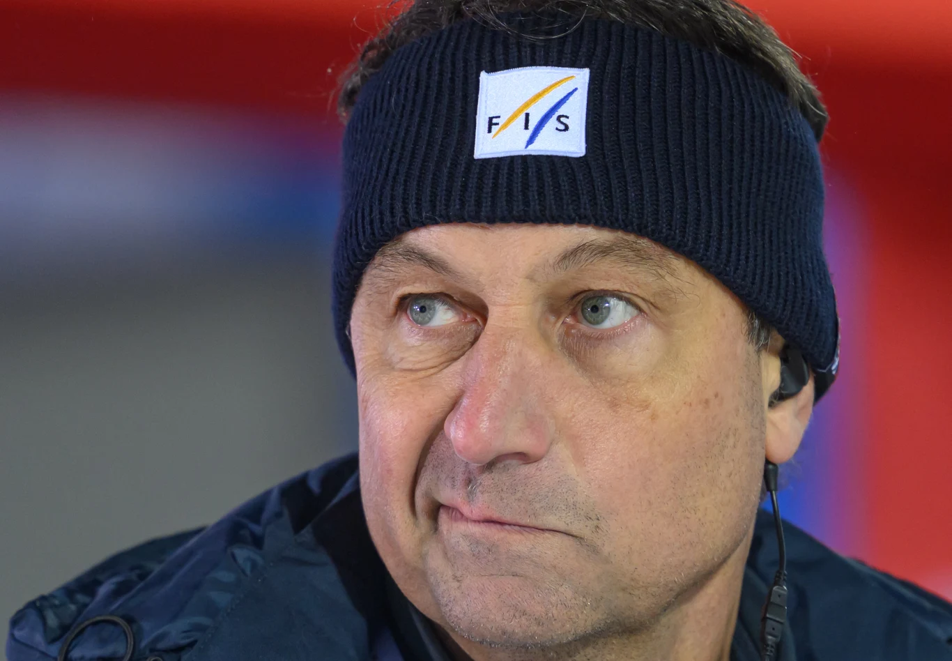 Mężczyzna w ciemnej opasce z logo FIS na głowie, ubrany w ciemną kurtkę, patrzy w bok z poważnym wyrazem twarzy, w uchu słuchawka.